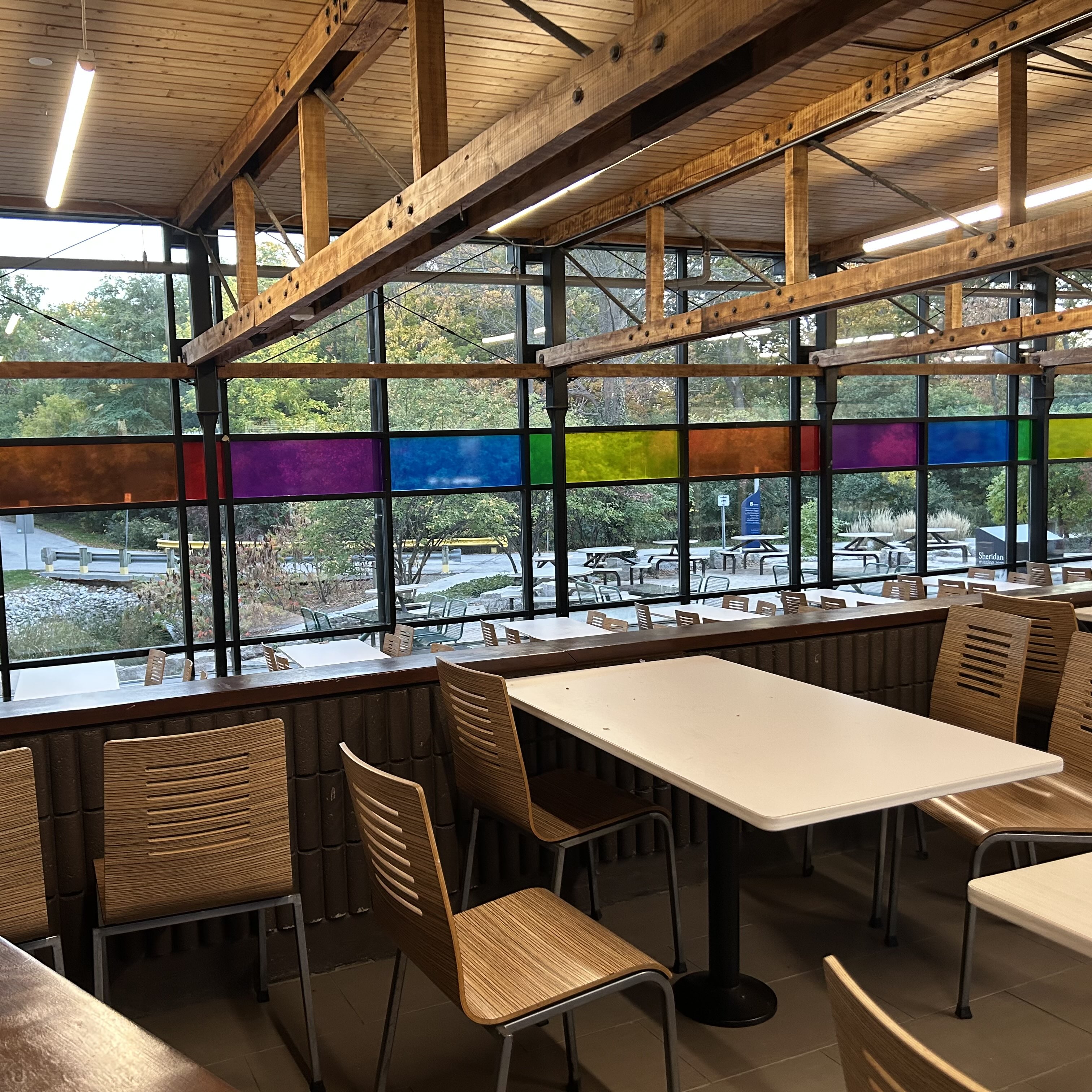 This is the cafetaria found in B wing, where students can sit down and order food. The chairs and tables seem to be made of wood and metal. The noticable long windows are made of glass, and certain glass panels that are coloured.